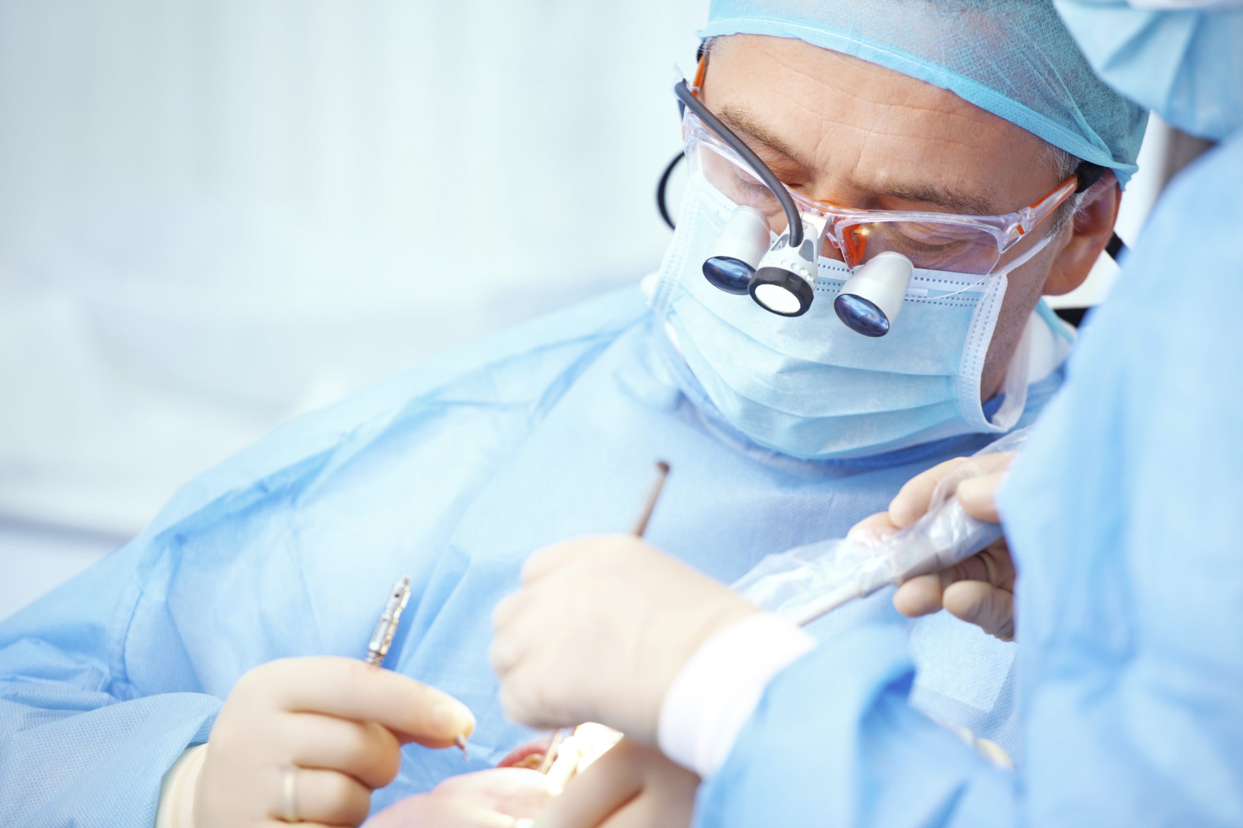 Dental professional using magnification loupes during a surgical dental procedure with chairside assistance in a clinical setting.